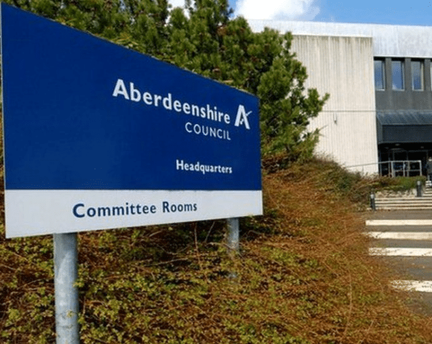 A sign reads "Aberdeenshire Council Headquarters, Committee Rooms" in front of a sleek, modern Aberdeenshire building surrounded by large steps and lush greenery. ©Mobysoft
