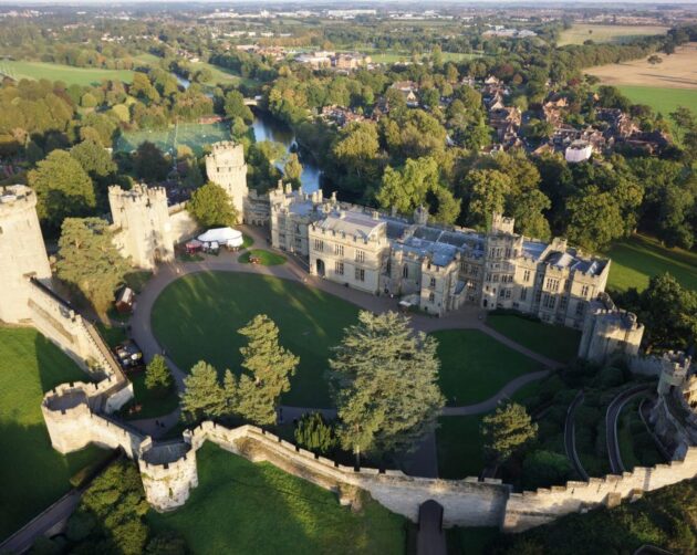 Aerial view of a large, historic stone castle surrounded by lush green trees and lawns. The castle features multiple turrets and walls, with a winding path leading up to it. As if part of an automated landscape, a small village and expansive scenery appear in the background. ©Mobysoft